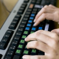 Foto van toetsenbord, op de lettertoetsen zijn groene en blauwe stickers geplakt. Twee handen typen op het toetsenbord.