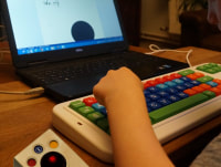 Foto van een laptop met daarvoor een kleurrijk en vergroot toetsenbord (Clevy) en een trackball. Een kinderhand typt op het toetsenbord.